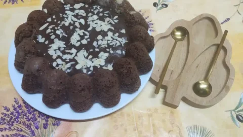 A chocolate cream cheese pound cake garnished with sliced almonds and chocolate glaze, served on a white plate with golden spoons on a wooden tray.