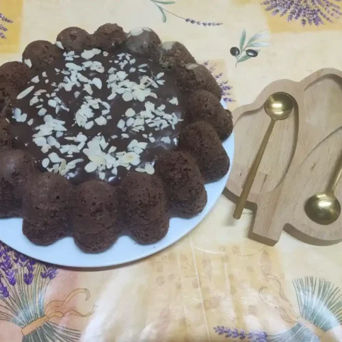 A chocolate cream cheese pound cake garnished with sliced almonds and chocolate glaze, served on a white plate with golden spoons on a wooden tray.