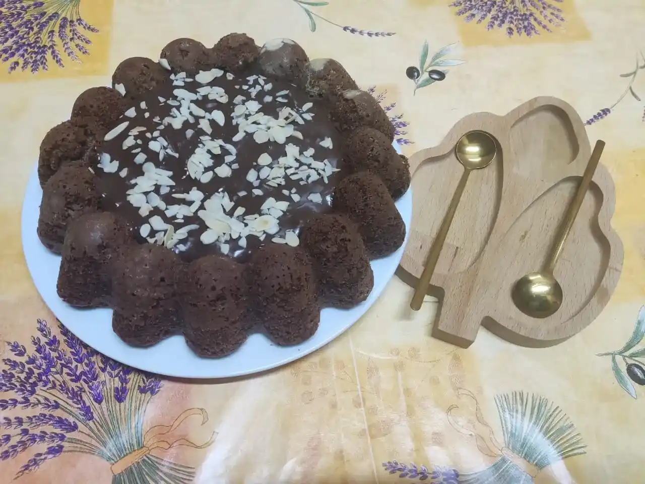 A chocolate cream cheese pound cake garnished with sliced almonds and chocolate glaze, served on a white plate with golden spoons on a wooden tray.