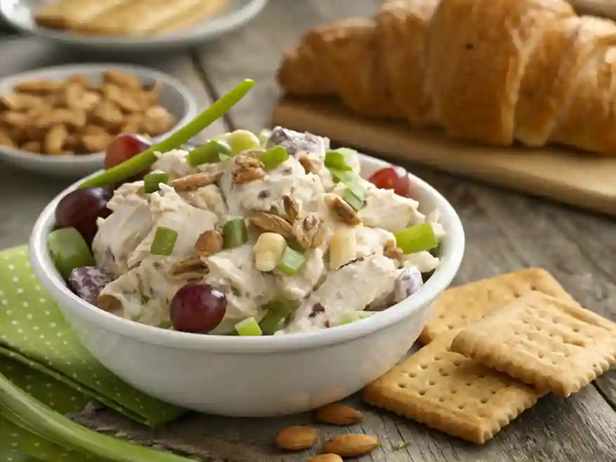 A creamy chicken salad with cream cheese, garnished with grapes, nuts, and green onions, served in a white bowl with a croissant and crackers on a rustic table.