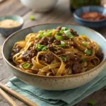 Realistic bowl of Mongolian Ground Beef Noodle with tender beef, noodles, and green onions on a rustic wooden table