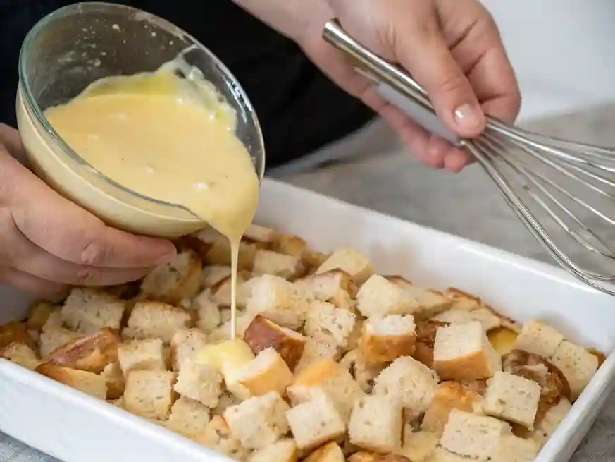 Hand pouring custard over sourdough bread cubes in a casserole dish